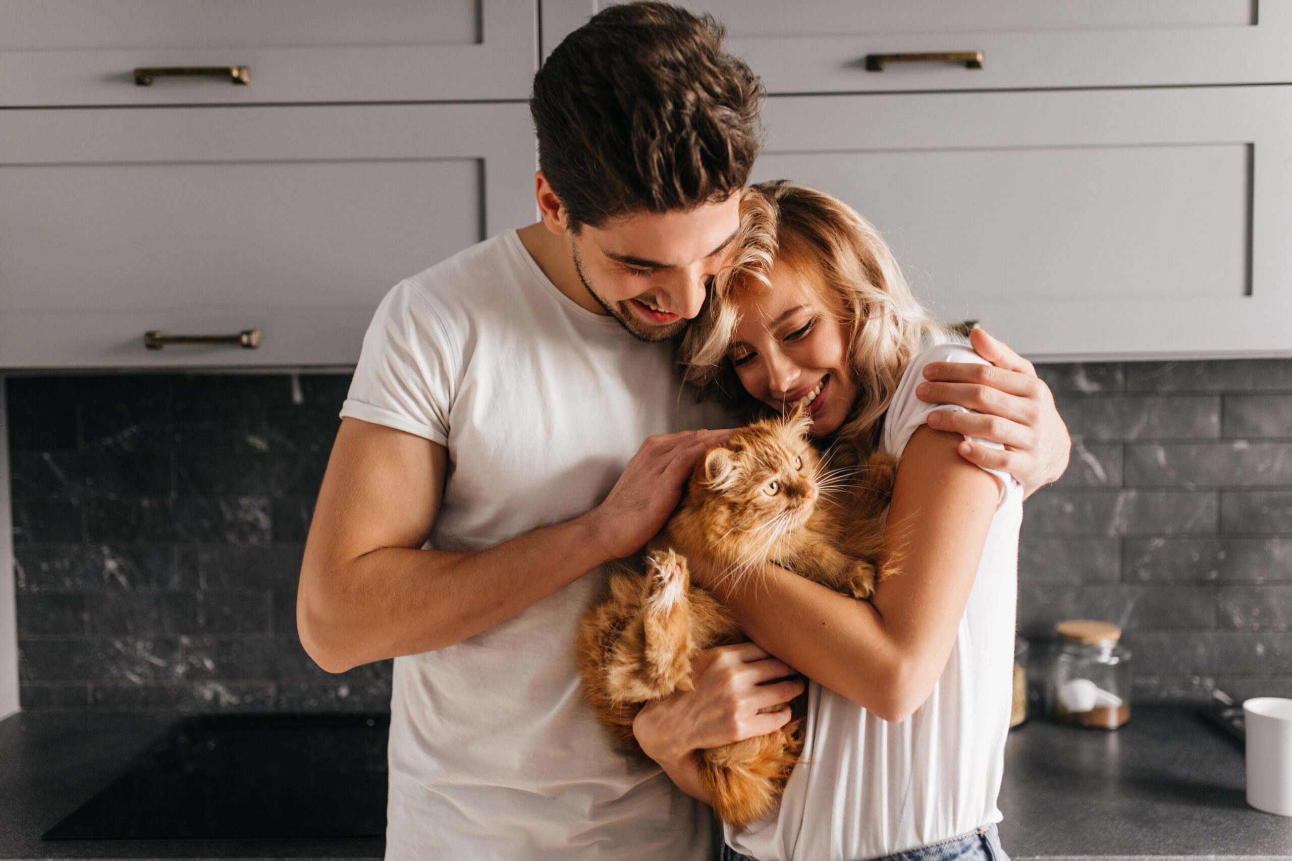 Brunette man looking at his cat and embracing wife. Indoor photo of happy family posing with pet.