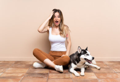 Young pretty woman with her husky dog sitting in the floor at indoors with surprise and shocked facial expression